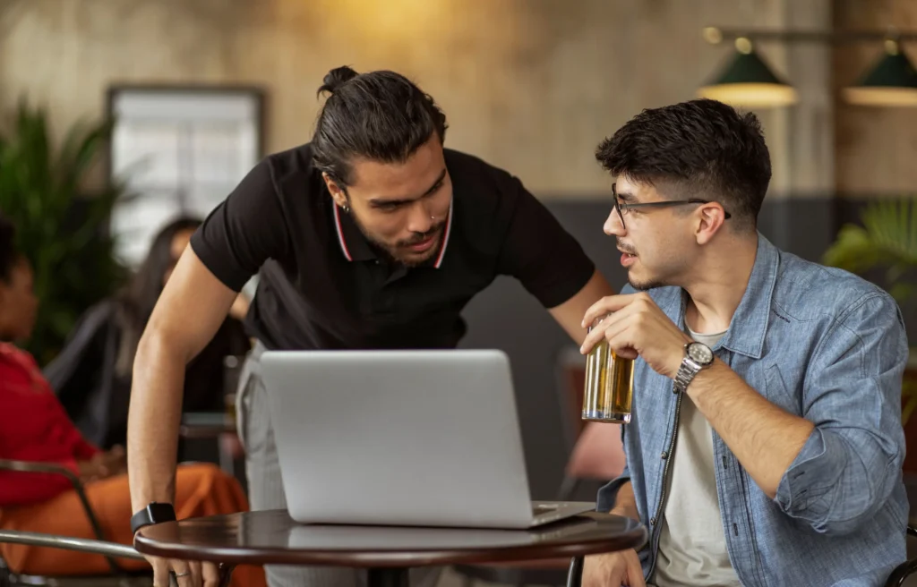 Two men collaborate in a cozy café setting while gathered around a laptop on a small round table. One man wearing glasses holds a drink while speaking, as his colleague in a black polo shirt leans forward to look at the screen and listen.