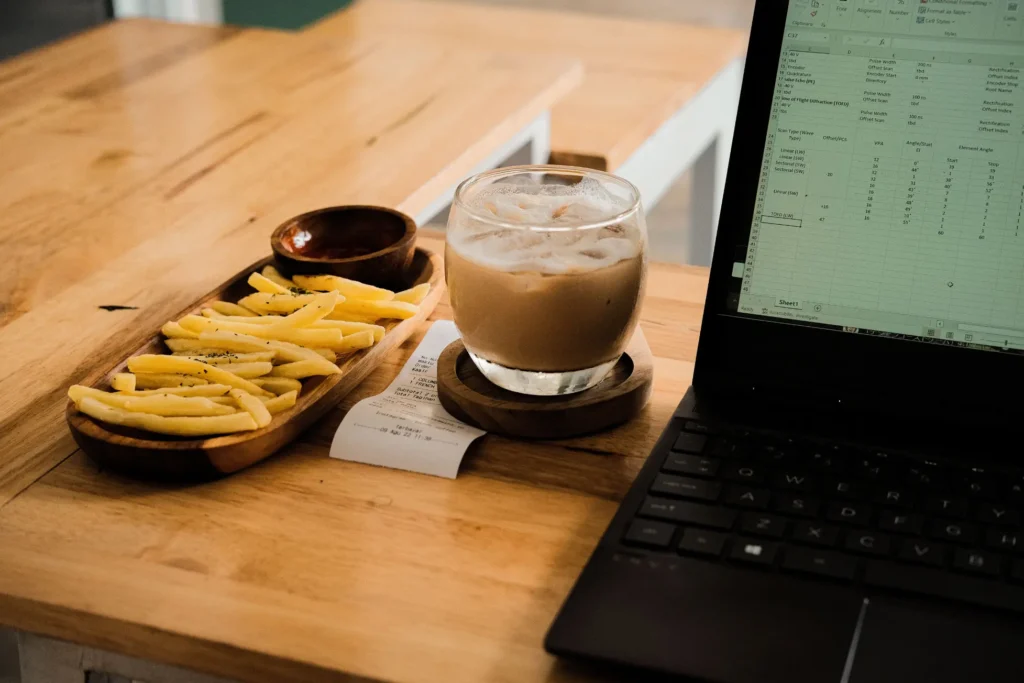 A glass of iced coffee resting on a wooden coaster sits on a table next to a tray of French fries and a small dipping bowl. Beside them, an open laptop displays a spreadsheet on its screen, along with a small printed receipt.