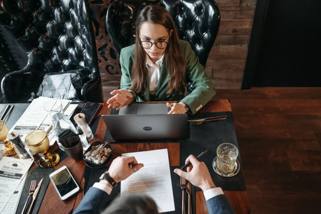 A woman wearing glasses and a green blazer speaks while looking at a laptop during a business meeting at a restaurant table. Across from her, a colleague holds a pen over a printed document surrounded by menus, glasses, and smartphones.