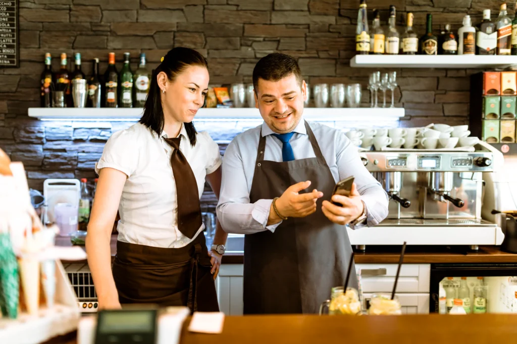 A male and female bartender stand behind a counter, smiling together as they look at a smartphone screen. In the background, shelves are lined with liquor bottles and a coffee machine.