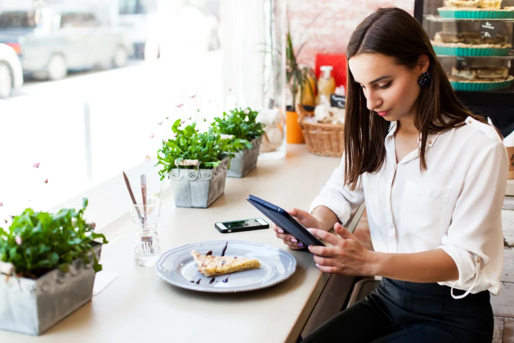 A young woman sits at a window counter in a cozy cafe, attentively looking at a tablet she holds in her hands. On the counter beside her are a plate with a slice of dessert, a smartphone, and potted green plants.