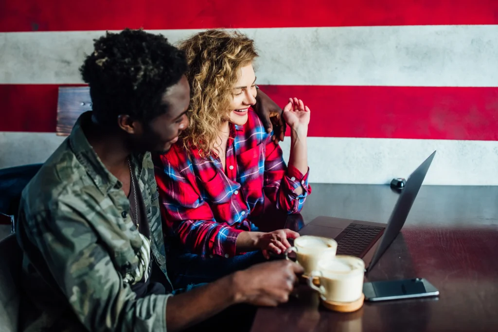 A smiling young couple sits together at a cafe table, looking intently at an open laptop screen. Two cups of coffee and a smartphone rest on the wooden surface in front of them against a bold red-and-white background.