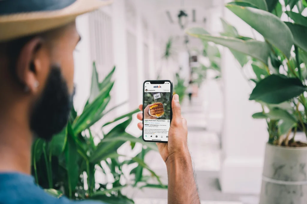 A man wearing a straw hat views a smartphone displaying a photo of a burger on the abillion app. He holds the phone outdoors along a bright, plant-lined white hallway.