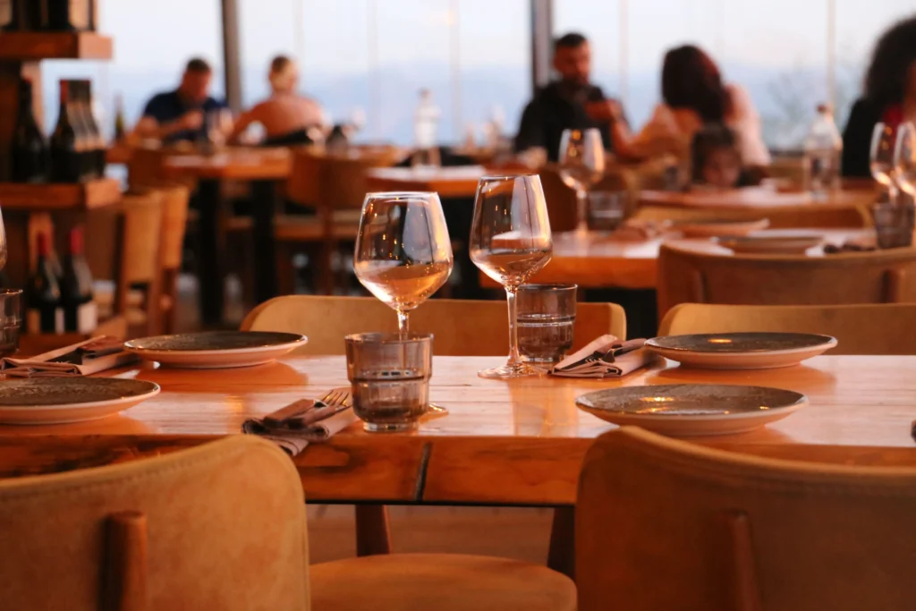 A close-up shot focuses on a wooden restaurant table set with wine glasses, water glasses, plates, and rolled napkins. In the softly blurred background, other diners sit at tables enjoying their meals near large glass windows.