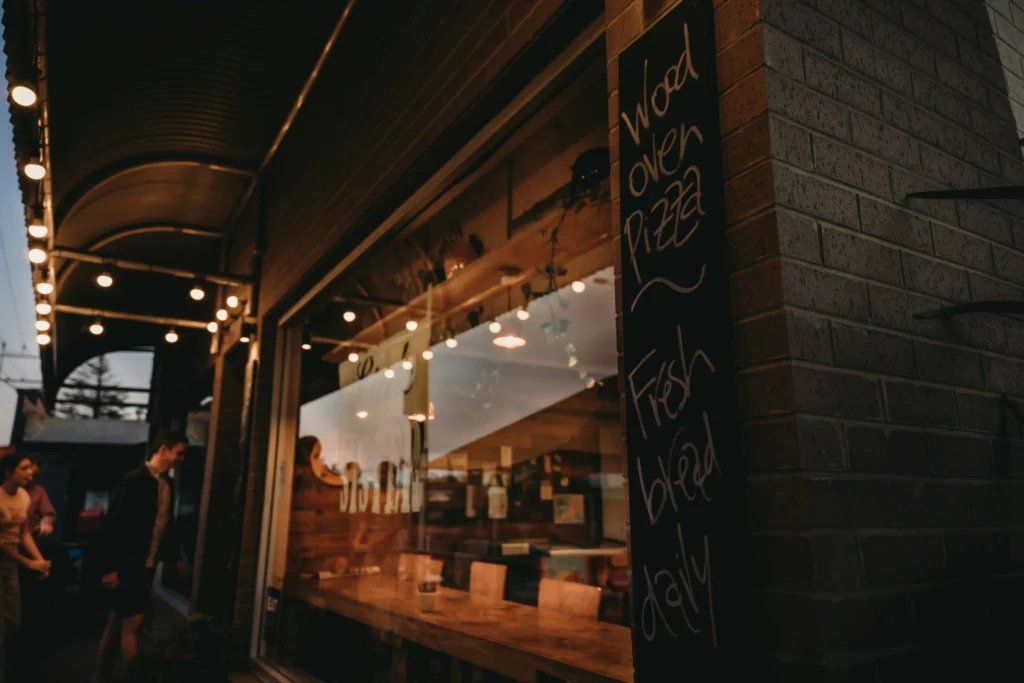 A cozy restaurant storefront lit with warm overhead string lights features a large glass window revealing indoor wooden seating. A tall chalkboard sign mounted on the exterior brick wall advertises "Wood oven Pizza" and "Fresh bread daily.”