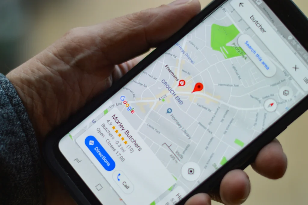 A close-up shot of a person holding a smartphone displaying Google Maps on its screen. The map shows a search for local butchers in the Crouch End area, highlighting a location card for Morley Butchers.