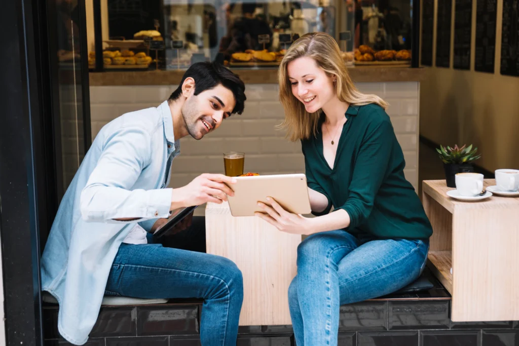 A smiling young man and woman sit together at an outdoor café table, looking at a digital tablet held by the woman. Coffee cups and pastries are visible in the background as the man points toward the screen.