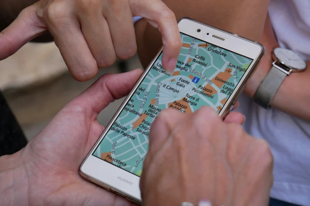 A close-up view shows a smartphone displaying a digital map of an Italian city held in hand. Multiple fingers point to specific locations on the screen as people collaborate to navigate their route.