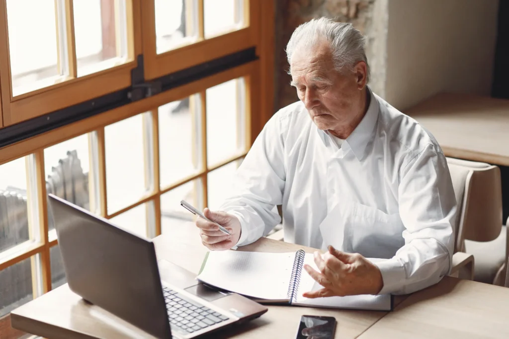 An elderly man with white hair wearing a white button-down shirt sits at a wooden table next to a large window. He holds a pen above an open spiral notebook while looking down intently, with an open laptop and smartphone placed beside him.