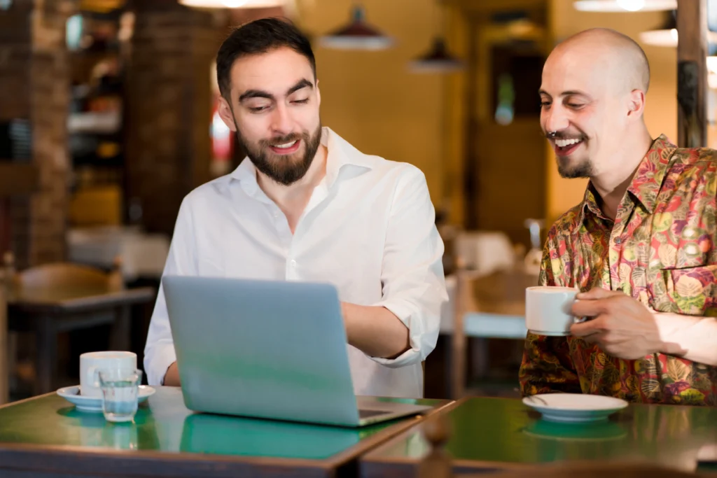 Two men sit together at a table in a cozy café with cups of coffee set in front of them. One man points toward an open laptop screen as his companion smiles and looks on.