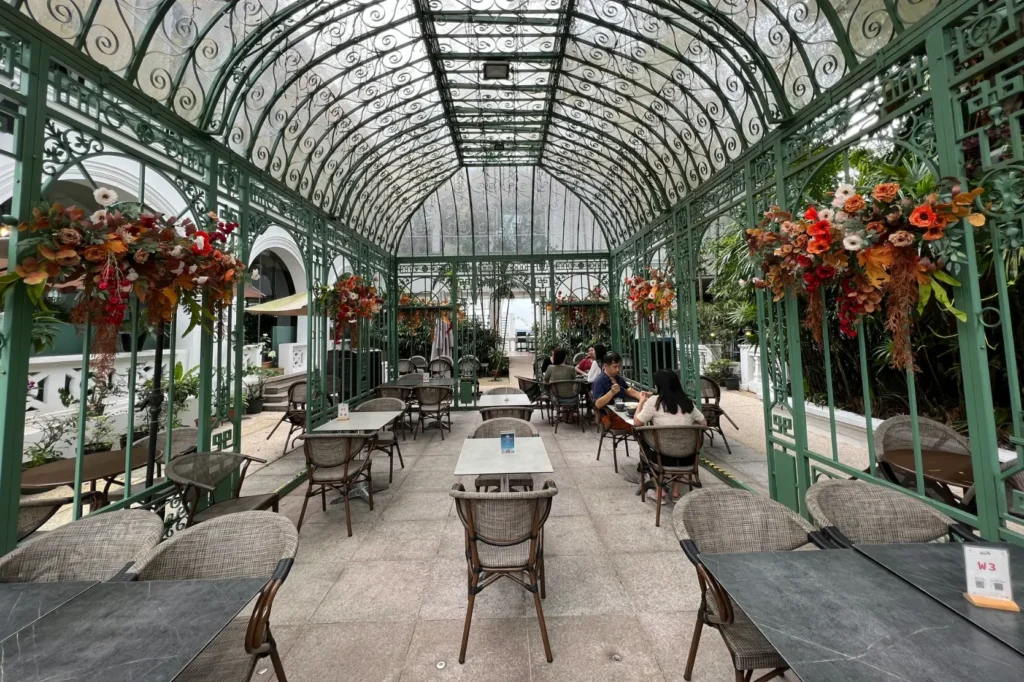 An elegant outdoor restaurant patio is enclosed within a green wrought-iron greenhouse structure featuring a vaulted glass ceiling. Warm autumn-colored floral arrangements adorn the ornate frame above small dining tables where several patrons sit.
