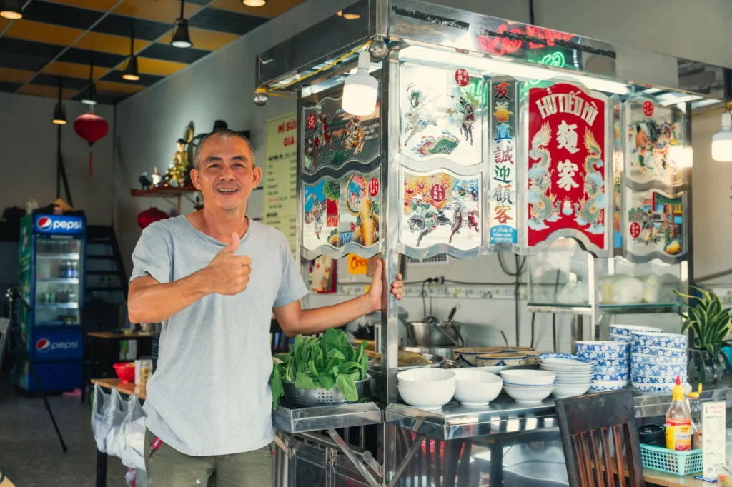 A smiling man wearing a grey t-shirt stands next to a stainless steel food cart and gives a thumbs-up gesture inside a restaurant setting. The noodle cart is decorated with colorful artwork, glass panels, and stacks of bowls ready for serving.