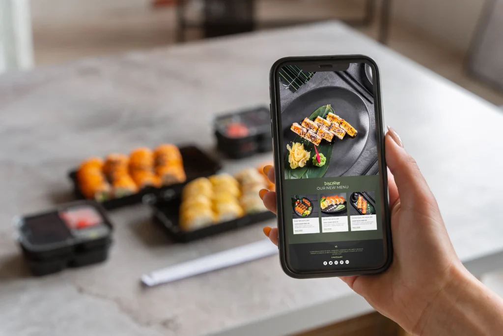 A person holds up a smartphone displaying a restaurant's sushi ordering app and menu. In the blurred background, several open takeout containers filled with fresh sushi rolls sit on a table.