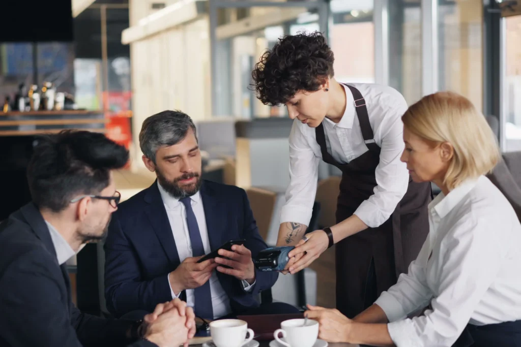 A server wearing an apron holds out a payment terminal to a group of business professionals seated at a table. One of the men prepares to pay using his smartphone while his colleagues look on.