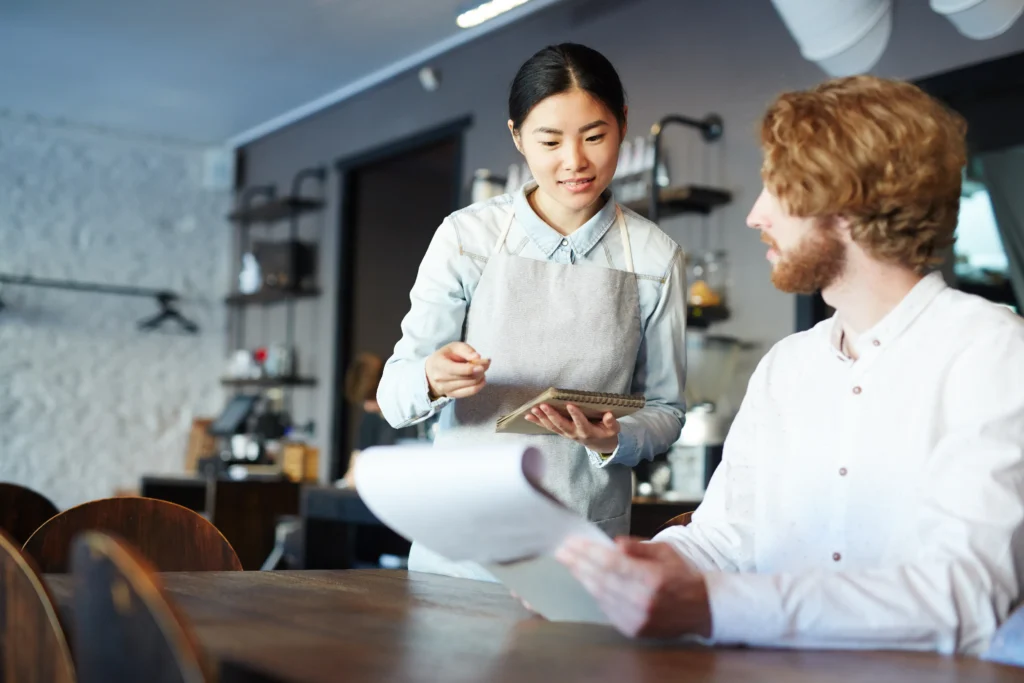 A waitress wearing a gray apron stands at a cafe table, holding a notepad and pen ready to take an order. A seated customer with reddish hair holds a menu as he speaks with her.