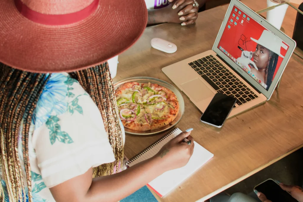 A person wearing a wide-brimmed red hat and braided hair works at a wooden table while taking notes next to a pizza. An open laptop displays a photo of a woman in a similar hat alongside a smartphone and a computer mouse.