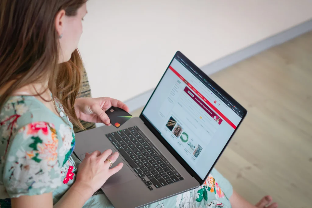 A woman wearing a floral dress sits with an open laptop on her lap, holding a credit card in her left hand. She appears to be browsing an online shopping website to complete a purchase.