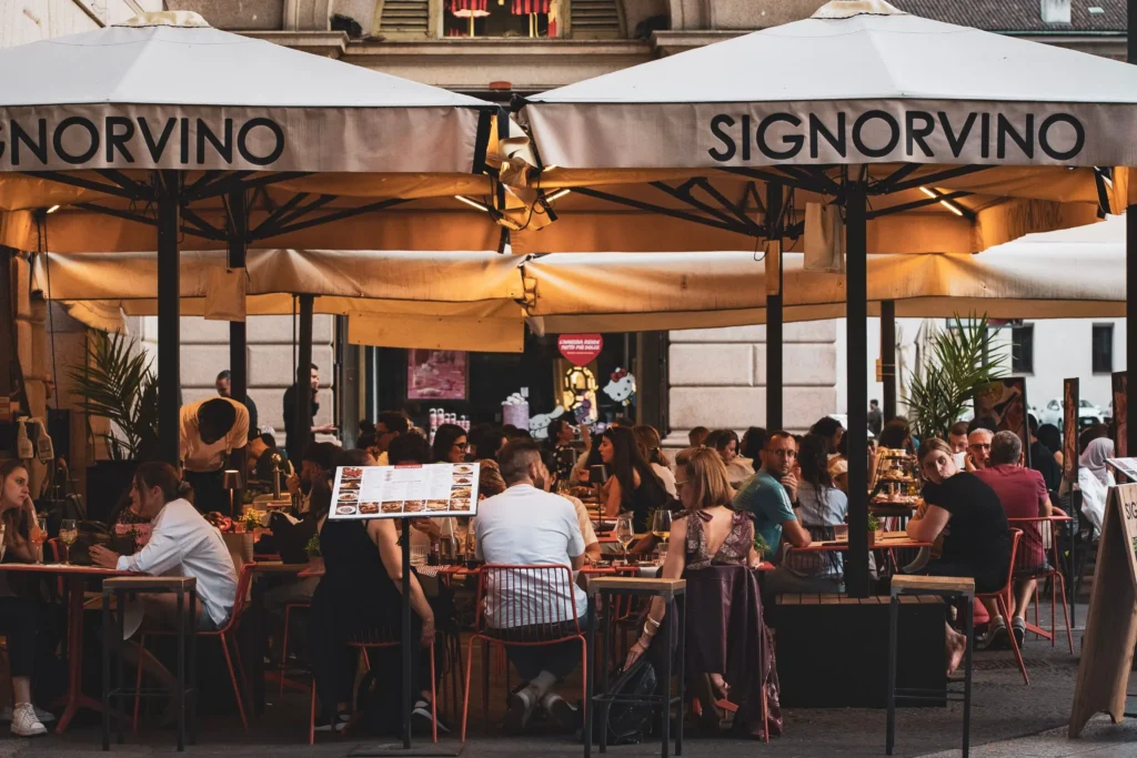 An outdoor patio at a Signorvino restaurant is bustling with patrons seated at tables under large white umbrellas. People are chatting, drinking wine, and dining in a warm, inviting setting.