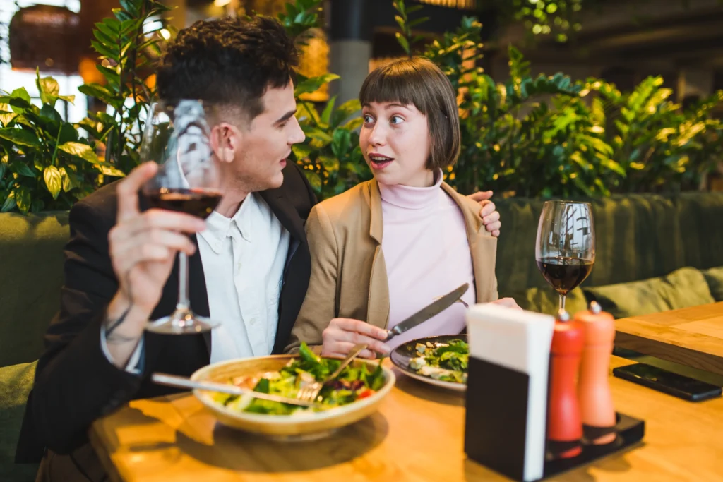 A young couple sits closely together at a restaurant table surrounded by lush greenery, enjoying red wine and salads. The man leans in holding a wine glass, while the woman looks at him with a pleasantly surprised expression.