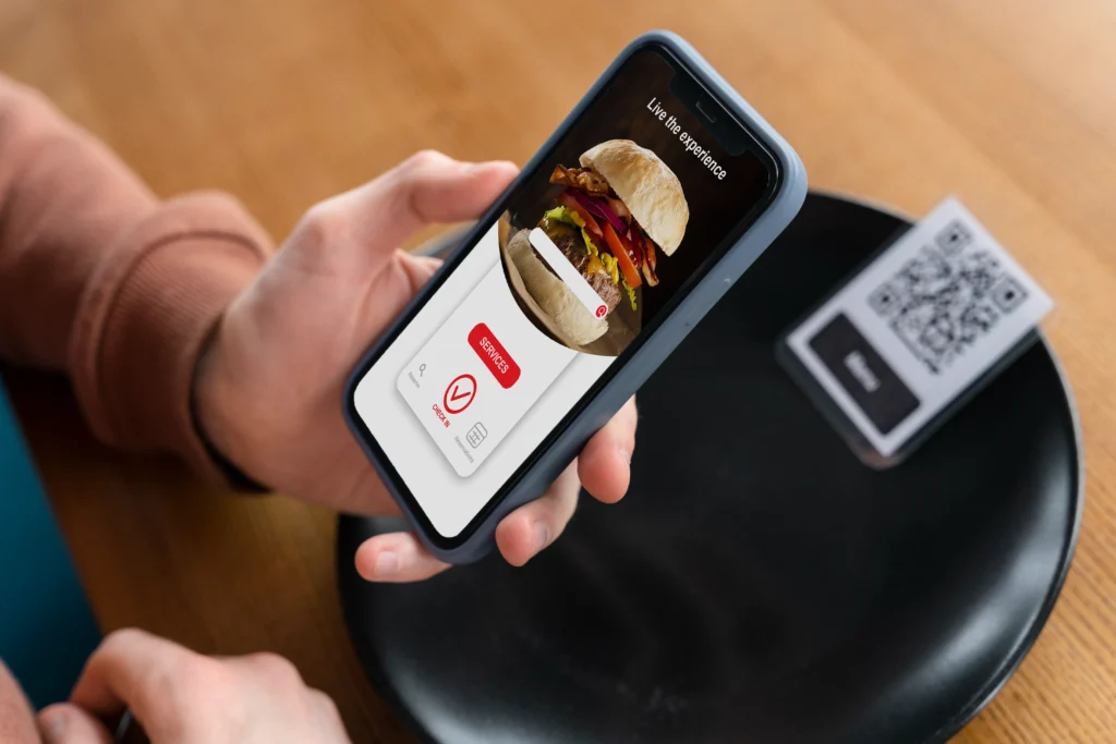 A close-up view of a person holding a smartphone that displays a digital restaurant menu featuring a burger. The phone rests above a wooden table with a black plate and a printed QR code.