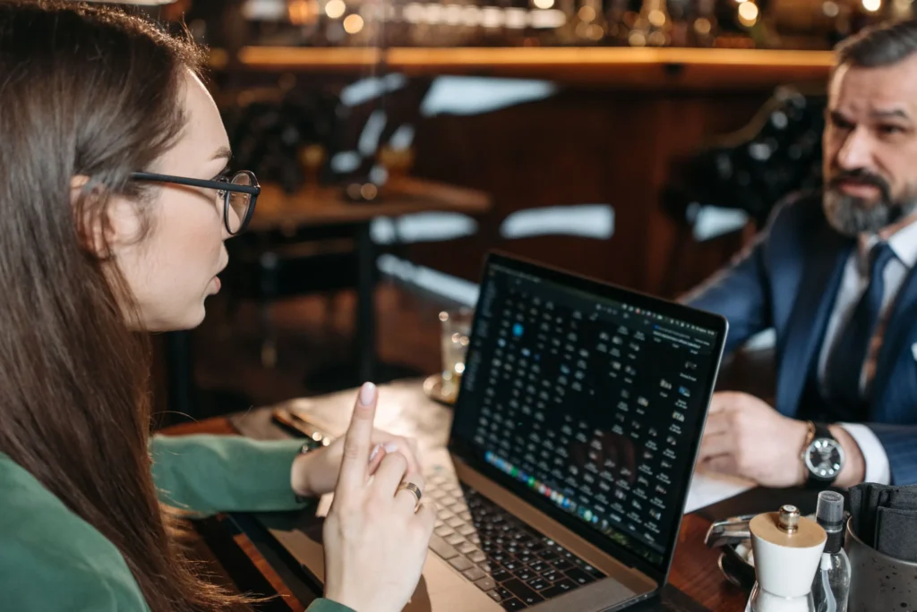 A woman wearing glasses gestures while discussing information displayed on her open laptop screen. Sitting across the table in a softly lit setting, a bearded man in a suit listens attentively to her presentation.