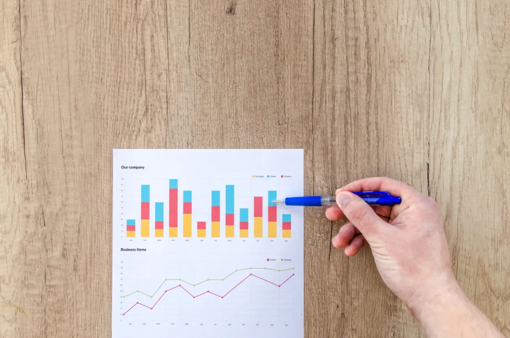 A person points a blue pen at a colorful stacked bar chart printed on a sheet of paper. The document sits on a wooden desk surface next to a line graph displaying business metrics.