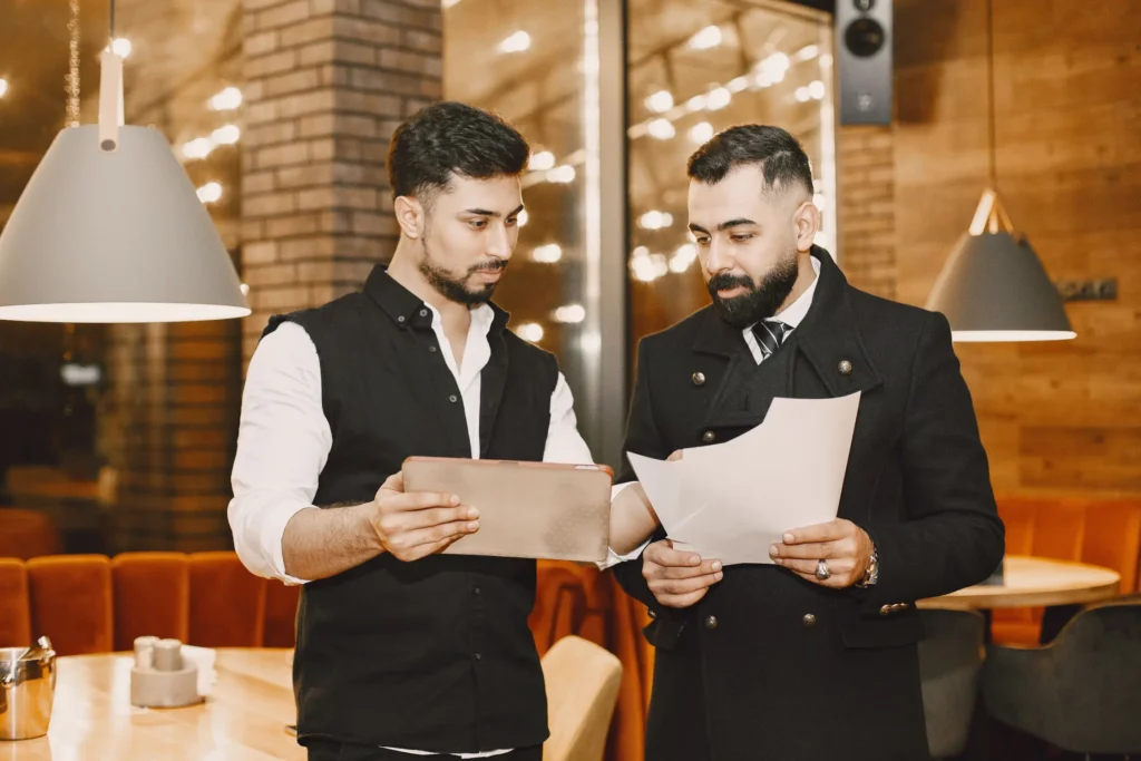 Two men stand side-by-side in a stylishly lit restaurant, discussing work documents. One holds a tablet while the other reviews printed papers in a double-breasted coat.