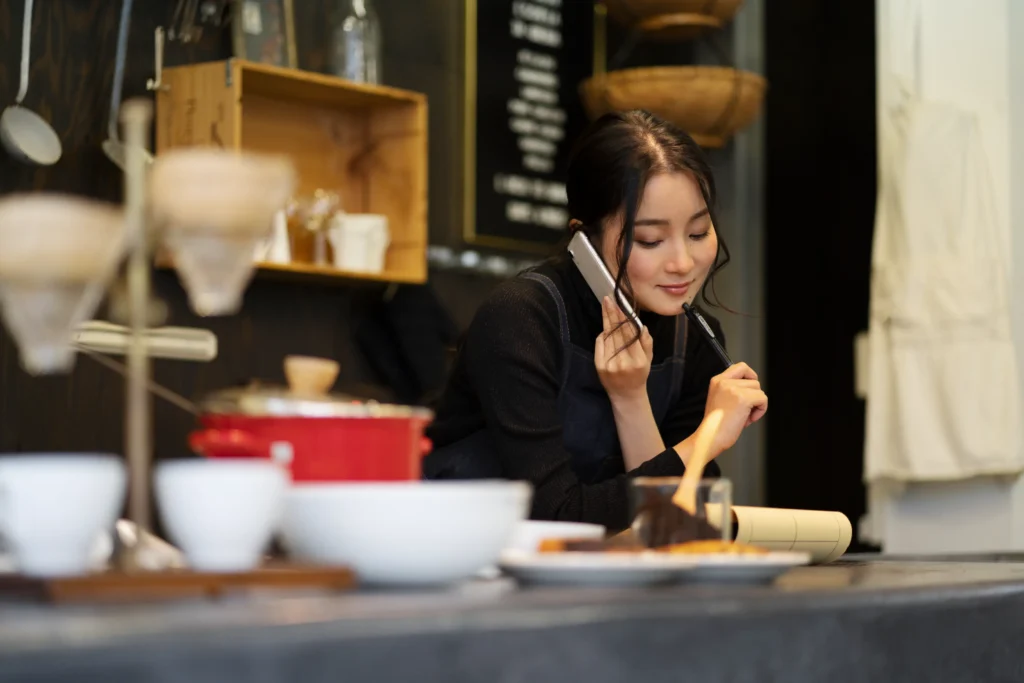 A woman in an apron talks on the phone while standing behind a cozy cafe counter. She holds a pen near her chin with a gentle smile, surrounded by coffee equipment and kitchenware.