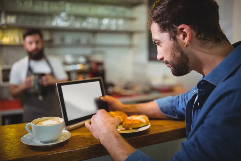 A bearded man in a blue shirt sits at a wooden cafe counter while looking closely at a digital tablet in his hands. Beside him sits a fresh cup of cappuccino with latte art and a plate of pastries, while a barista stands blurred in the background.