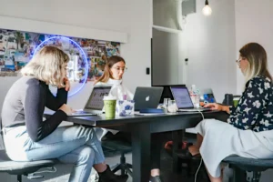 Three women sit around a large black conference table in a bright, modern office space working on their laptops. Behind them, a circular neon light illuminates a photo mood board mounted on the wall.