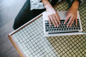 An overhead view shows a person wearing a denim jacket typing on a silver laptop. The laptop rests on a patterned glass-tiled table with a wooden frame.