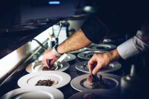 A chef carefully places garnishes onto dishes lined up under the pass light in a dimly lit restaurant kitchen. The close-up view highlights the precise hands-on plating process across multiple white plates.