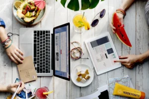 Top-down view of people sitting at a white wooden table using a laptop and a tablet while planning a vacation. The table is filled with summer essentials, including tropical fruit, drinks, sunglasses, a postcard, and sunscreen.