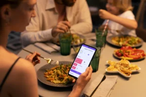 A woman holds a fork over a pasta plate while using a smartphone app to control ambient lighting settings. Her family sits around the warm dining table enjoying their dinner in the background.