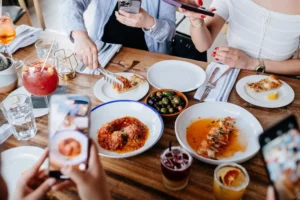 A top-down view of a wooden dining table filled with colorful dishes like meatballs, pasta, olives, and cocktails. Friends gather around the table, serving food and holding up smartphones to take photos of their meal.