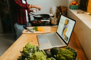 A person in a red plaid shirt seasons food cooking on the stove while following a recipe on an open laptop on the kitchen counter. Fresh vegetables and prepped ingredients surround the workspace.