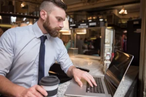 A man with a beard, light blue shirt, and tie works on a laptop at a café counter. He holds a takeaway coffee cup while intently focusing on his screen.