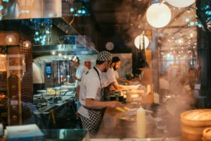 Chefs work actively in a bustling restaurant kitchen filled with warm lighting and steam from fresh cooking. Seen through a glass window, they prepare dishes behind a counter lined with squeeze bottles and kitchen equipment.