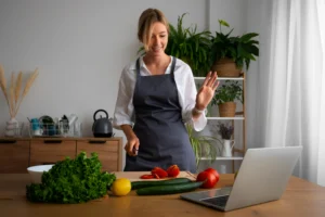 A woman wearing an apron smiles and waves at a laptop screen while standing in a bright kitchen. Fresh vegetables are laid out on the wooden table in front of her as she prepares food.