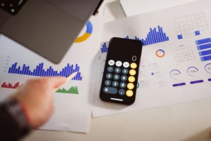 Top-down view of a workspace with printed financial reports, bar charts, and a smartphone displaying a calculator app in the center. A person's hand is visible on the left, pointing a pen toward a blue bar graph.