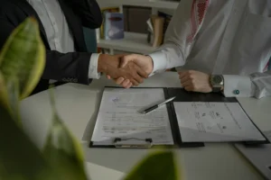Two professional men shake hands across a desk over signed business documents and a silver pen. One man wears a dark suit jacket while the other is dressed in a traditional white thobe and ghutra.