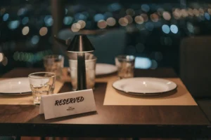 A dimly lit dining table is set with empty plates, drinking glasses, a small lamp, and a white card that reads "RESERVED." In the background, blurry bokeh lights create a romantic nighttime view of the city skyline.