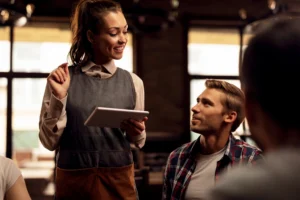 A smiling waitress holding a tablet takes an order from seated customers inside a dimly lit café. She interacts warmly with a man sitting at the table while gesturing with her hand.
