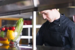 A chef dressed in a black jacket focuses intently on preparing a dish in a commercial kitchen line. A bowl filled with fresh produce, including tomatoes, lemons, and romaine lettuce, sits in the foreground.