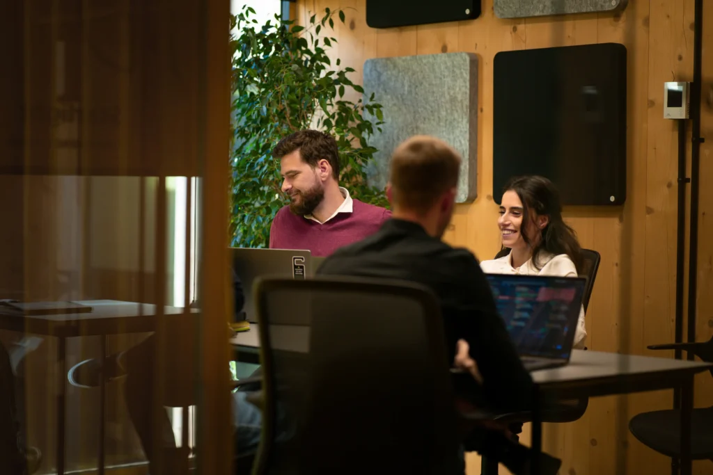 Three colleagues sit around a conference table working on open laptops in a modern office with light wood walls. One woman smiles in conversation while a bearded man beside her focuses on his computer screen.