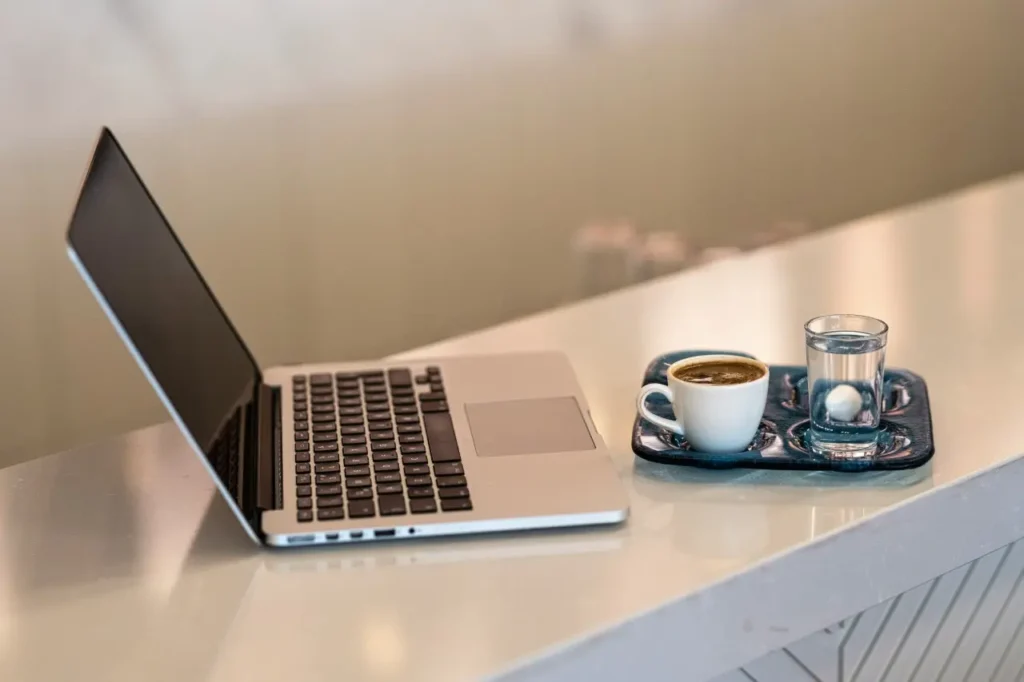 An open laptop sits on a sleek, light-colored counter next to a serving tray with a small cup of espresso. Beside the coffee, a small glass of water with a white sweet or ice cube completes the minimal workspace setup.