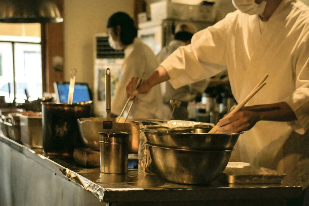 Chefs wearing white jackets and face masks prepare food along a stainless steel kitchen counter. One chef in the foreground uses large wooden chopsticks over mixing bowls while another works with utensils at a stove.