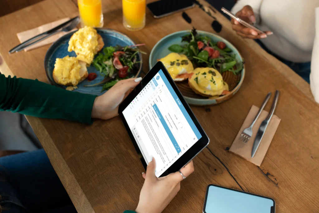 An overhead view of a person using a tablet to read an email at a restaurant table with breakfast plates and orange juice. Another person nearby holds a smartphone, ignoring their untouched meal.