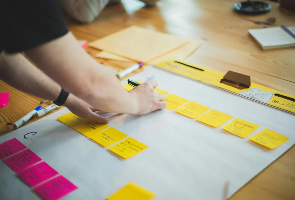 A person writes on a yellow sticky note placed alongside several pink and yellow notes organized on a large white paper easel pad over a wooden table. The surrounding workspace is set up for a brainstorming session, complete with markers, notebooks, and folders.