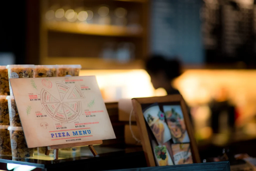 An illustrated pizza menu sits on a small wooden display easel on a cafe counter alongside stacked plastic containers. The background is softly blurred, showing warm indoor lighting and a coffee shop interior.